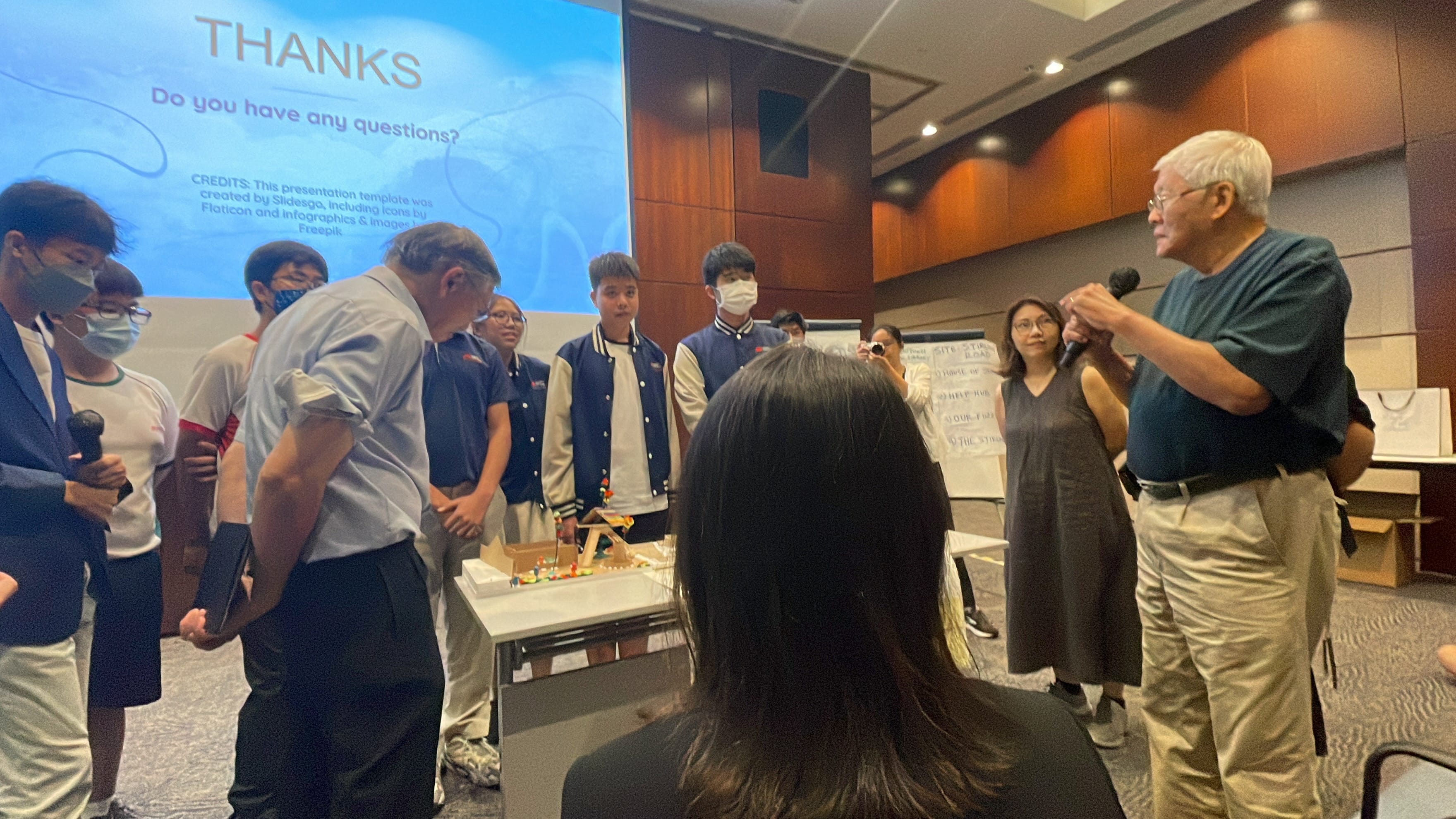 Students and adults gather around a table with a model during a Q&A session, with a "Thanks" slide projected behind them.